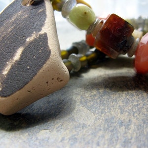 May include: A close-up of a necklace featuring a mix of beads, including a large, brown, textured, heart-shaped pendant. The beads are made from glass, stone, and metal. The necklace is displayed on a grey surface.