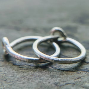 Könnte beinhalten: Zwei silberne Creolen, eine etwas größer als die andere, liegen auf einer strukturierten grauen Oberfläche. Die Ohrringe sind aus dünnem, gehämmertem Metall.