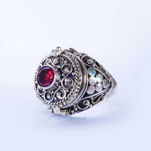 Puede incluir: Un anillo de plata con un dise&ntilde;o de filigrana ornamentado. El anillo presenta una gema roja grande y redonda en el centro y un compartimento con bisagras. El anillo est&aacute; marcado con el texto "Katybeads".