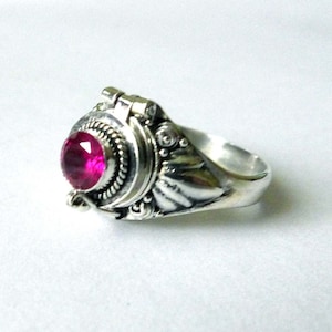Puede incluir: Un anillo de plata con una gran gema magenta redonda. El anillo tiene un diseño ornamentado con detalles de metal alrededor de la piedra y en la banda. El anillo está sobre un fondo blanco.