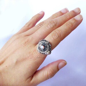 Puede incluir: Anillo de plata ornamentado con una gema roja central rodeada de intrincados filigranas y peque&ntilde;os detalles blancos. El anillo se muestra en una mano, resaltando su dise&ntilde;o detallado. Una pieza de joyer&iacute;a &uacute;nica.