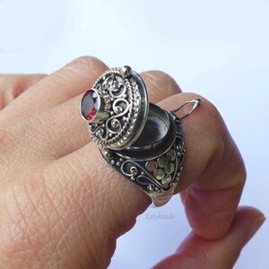 Puede incluir: Un anillo de plata con un dise&ntilde;o de medall&oacute;n ornamentado. El anillo presenta una tapa con bisagras con una piedra preciosa roja redonda. El anillo est&aacute; abierto, revelando un peque&ntilde;o compartimento. El anillo se lleva en un dedo.