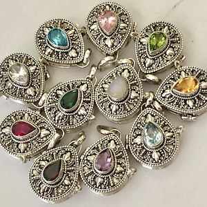 May include: A collection of silver-toned teardrop-shaped pendants with various colored gemstones. Each pendant features an ornate, detailed design around the central stone, including pink, blue, green, and yellow gems. The pendants are arranged on a light-colored surface.