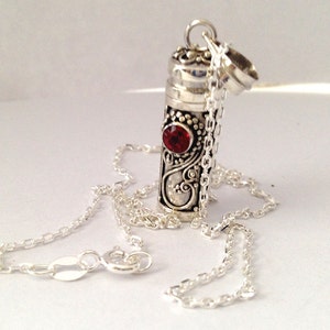 Puede incluir: Un collar con colgante plateado con un relicario cilíndrico, detallado con patrones ornamentados y una gema roja. El relicario cuelga de una fina cadena, con la parte superior abierta. El diseño incorpora motivos arremolinados y un cierre seguro.