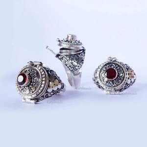 Puede incluir: Tres anillos de plata ornamentados con un compartimento con bisagras y una gema roja. Los anillos presentan intrincados dise&ntilde;os de volutas y flores. Un anillo est&aacute; abierto, revelando el interior. Los anillos se muestran sobre un fondo blanco.