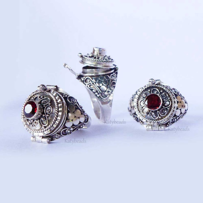 Puede incluir: Tres anillos de plata ornamentados con un compartimento con bisagras y una gema roja. Los anillos presentan intrincados dise&ntilde;os de volutas y flores. Un anillo est&aacute; abierto, revelando el interior. Los anillos se muestran sobre un fondo blanco.