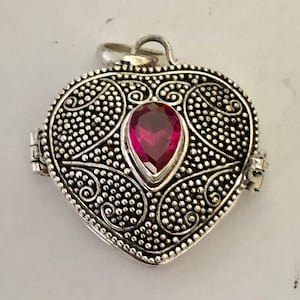 May include: A silver-colored heart-shaped pendant with a hinged opening. The pendant features a central, teardrop-shaped, deep red gemstone. The surface is intricately detailed with a dotted pattern and swirling designs.