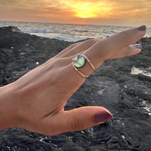 Pode incluir: Um anel dourado com um centro redondo de concha de abalone iridescente. A banda do anel tem um design texturizado. O fundo apresenta um pôr do sol sobre o oceano.