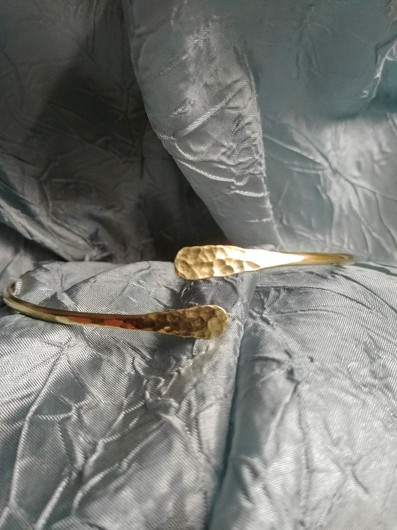 May include: A hammered gold bangle bracelet with a slightly curved shape. The bracelet is made of a thick, textured metal.
