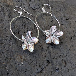 May include: A pair of silver hoop earrings, each adorned with a flower-shaped pendant. The flower pendants have a pearlescent, iridescent finish, with shades of white, pink, and gold. The earrings are displayed on a dark, textured surface.