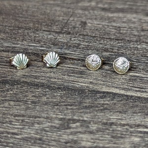 Puede incluir: Cuatro pendientes de color dorado sobre una superficie de madera. Dos pendientes tienen forma de concha y dos son redondos con un diseño floral.