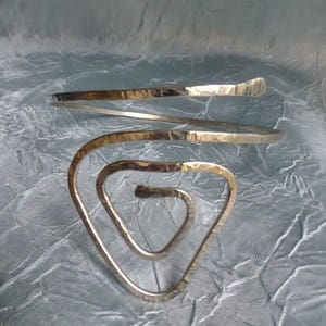Puede incluir: Un brazalete de metal martillado plateado con un diseño en espiral. El brazalete es abierto y ajustable.