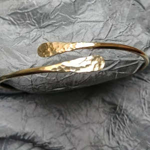 May include: A gold hammered metal bangle bracelet with a simple, open design. The bracelet is made of a single piece of metal that is bent into a circle and then hammered to create a textured surface.