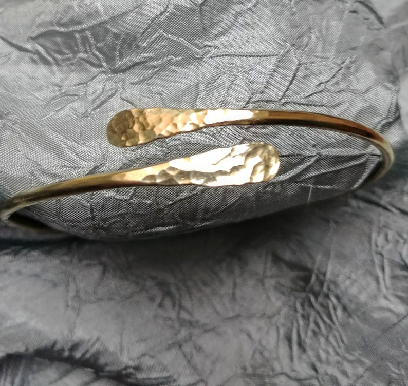 May include: A gold hammered metal bangle bracelet with a simple, open design. The bracelet is made of a single piece of metal that is bent into a circle and then hammered to create a textured surface.