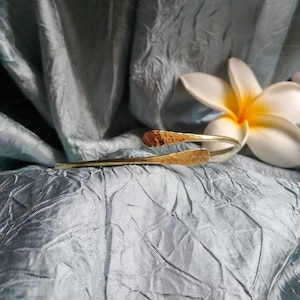 May include: A gold hammered bangle bracelet with a simple, open design. The bracelet is lying on a blue textured fabric.