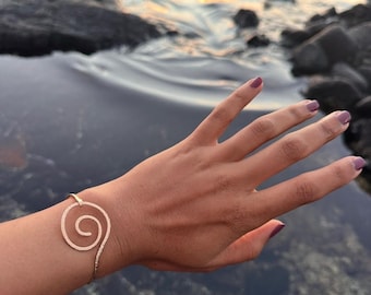 Puede incluir: Un brazalete de metal en tono oro rosa con un diseño de espiral prominente en la parte frontal. La pulsera presenta una banda delgada y pulida que se curva en un patrón de remolino circular texturizado, formando un bucle abierto en la muñeca.