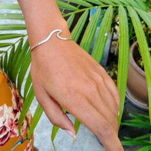 Puede incluir: Una pulsera de plata en forma de ola en la muñeca de una persona. La pulsera está hecha de metal y tiene una textura martillada.