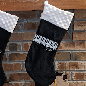 May include: Two black Christmas stockings with white faux fur cuffs. The stocking on the right has a white piano keyboard design with the word "Cursive" printed below it.