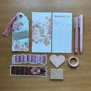 May include: A stationery set including a floral bookmark with a pink tassel, a floral card with a quote, a white reading list tracker, two light pink pens, a purple book sticker, a library sticker, a heart-shaped tag, and a roll of tape.