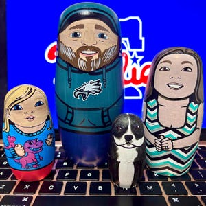 May include: Four hand-painted wooden nesting dolls, or matryoshka dolls, depicting a family. The largest doll is a man with a beard wearing a green hoodie with an eagle logo. The next doll is a woman with long brown hair wearing a blue and white striped dress. The third doll is a girl with blonde hair wearing a blue shirt with a pink dinosaur. The smallest doll is a black and white dog.