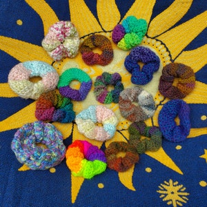 May include: A collection of 16 colorful crocheted hair scrunchies in various patterns and colors, including rainbow, pink, blue, green, and brown.
