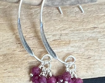 Pode incluir: Brincos de argola prateados com textura martelada. Cada brinco apresenta um aglomerado de pequenas contas redondas de cor vermelho escuro. Um par de pequenos brincos de botão prateados também é visível.