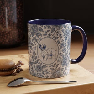 May include: A ceramic mug with a blue interior and handle, featuring a floral pattern in blue and white. The mug has a central oval with a detailed illustration of a dog. A spoon and a pastry are on a wooden surface.