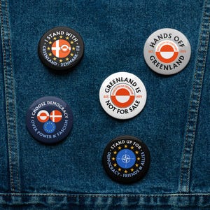 Puede incluir: Una colección de botones redondos con varios diseños y texto. Los botones presentan la bandera de Groenlandia, la bandera de la UE y frases como "I Stand With Greenland-Denmark-EU" y "Hands Off Greenland."