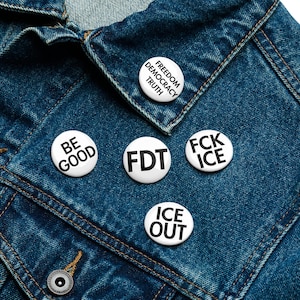 Puede incluir: Primer plano de una chaqueta vaquera azul oscuro adornada con cinco botones blancos. Los botones muestran textos negros: "Freedom Democracy Truth", "Be Good", "FDT", "FCK ICE" y "ICE OUT".