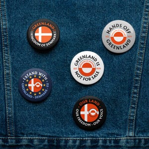 May include: A selection of circular badges displaying Greenlandic flag designs and text. The badges feature the Greenland flag, the words "Greenland," "Kingdom of Denmark," and phrases such as "Hands Off" and "Not For Sale."