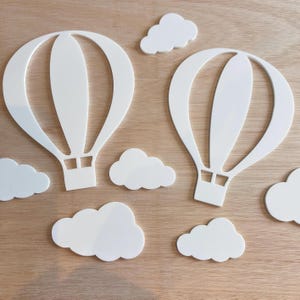 Puede incluir: Recortes blancos de globos aerostáticos y nubes. Los globos tienen una cesta en la parte inferior y están rodeados de formas de nubes esponjosas. Los recortes están dispuestos sobre una superficie de madera marrón claro.