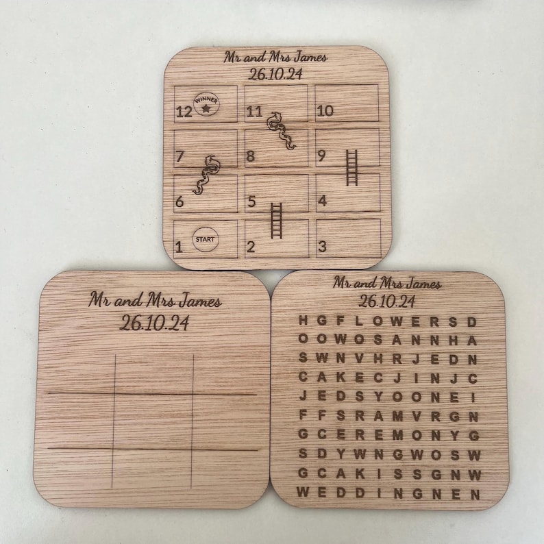 May include: Three wooden game boards with the text "Mr and Mrs James 26.10.24" on each. The first board has a game board with numbers 1-12 and images of ladders and snakes. The second board is a blank game board with lines for players to draw their own game. The third board has a word search puzzle with the words "HGFLOWERSD", "OOWOSANNHA", "SWNVHRJEDN", "CAKECJINJC", "JEDSYOONEI", "FFSRAMVRGN", "GCEREMONYG", "SDYWNGWOSW", "GCAKISSGNW", and "WEDDINGNEN".