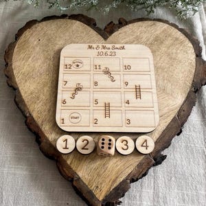 May include: A wooden heart-shaped game board with a snakes and ladders game printed on it. The board is personalized with the text "Mr & Mrs Smith 10.6.23". There are four wooden dice with numbers 1, 2, 3, and 4 printed on them.