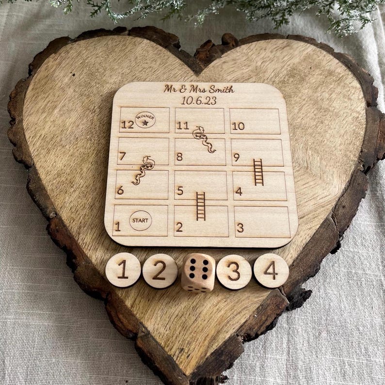 May include: A wooden heart-shaped game board with a snakes and ladders game printed on it. The board is personalized with the text "Mr & Mrs Smith 10.6.23". There are four wooden dice with numbers 1, 2, 3, and 4 printed on them.