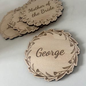 May include: Three round wooden signs with a floral wreath design and engraved text. The top sign reads "Mother of the Bride". The bottom sign reads "George".