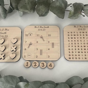 May include: A wooden game set with two boards. One board has a tic-tac-toe game with hearts and X's. The other board has a game with a ladder and a snake. The boards are labeled with the text "Mr. & Mrs. Smith" and the date "10.6.23". There are four wooden game pieces with the numbers 1, 2, 3, and 4.