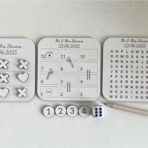 May include: A wedding game set with a tic-tac-toe board, a word search, a numbered game board, and a dice. The game boards are white with black text and graphics. The dice is white with black dots. The game set is perfect for a wedding reception or party.