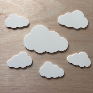 May include: Six white cloud-shaped decorations of varying sizes are arranged on a light wood-grain surface. The clouds have a smooth, solid appearance, and are likely made of a durable material. These decorative clouds could be used for nursery decor or craft projects.