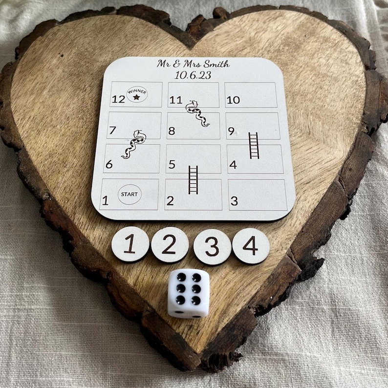 May include: A wooden heart-shaped board with a white game board featuring a snakes and ladders game. The game board has the text "Mr & Mrs Smith 10.6.23" at the top. There are four white game pieces with black numbers 1, 2, 3, and 4. A white die with black dots is in the center of the board.