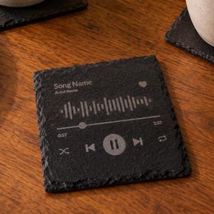 May include: A square black slate coaster featuring a laser-engraved music player interface. The design includes the text Song Name and Artist Name, a waveform graphic, a heart icon, a progress bar, and playback controls including pause and skip.
