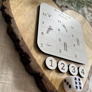 May include: A wooden heart-shaped board with a silver game board featuring a snakes and ladders game. The game board has numbers 1 through 12, with the words "Start" and "Winner" printed on the board. The board is surrounded by white numbered game pieces and a white die with black dots.