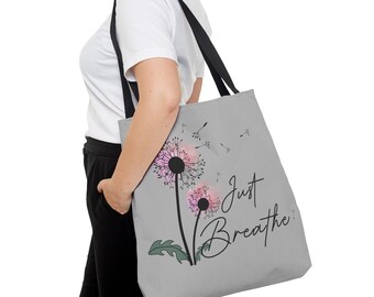 May include: A gray tote bag with black straps features a watercolor dandelion design with pink and green accents. The text "Just Breathe" is printed in black cursive lettering.