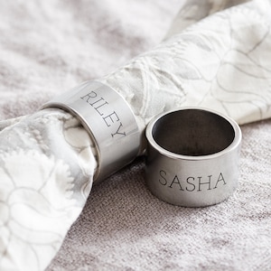 May include: Two silver napkin rings with the names "Riley" and "Sasha" engraved on them. The rings are on a light gray fabric surface.
