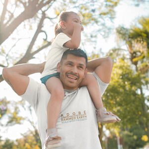 Op de afbeelding: Een man draagt een kind op zijn schouders. Hij draagt een wit T-shirt met de tekst "ADOBE COMPRESSED EARTH BLOCK RAMMED EARTH BUILD WITH THE EARTH" erop gedrukt. Het kind draagt een wit T-shirt en heeft zijn armen om de nek van de man.