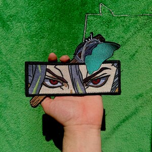 Peut inclure: Un écusson rectangulaire représentant une illustration de style anime des yeux et des cheveux d'un personnage. Les yeux sont rouges et les cheveux gris et verts. Un papillon aux ailes turquoise est attaché au dessus.
