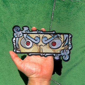 Peut inclure: Écusson brodé représentant un visage stylisé avec des yeux rouges intenses, sur un fond beige et doré. L'écusson a une forme rectangulaire avec une bordure bleue décorative. L'écusson est tenu dans une main sur un fond vert.