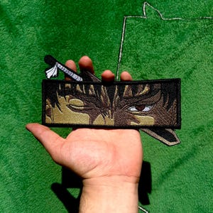 Peut inclure: Écusson brodé représentant un visage de personnage aux tons marron et or, et une épée. L'écusson est rectangulaire avec une bordure noire. L'écusson est tenu dans une main sur un fond vert.