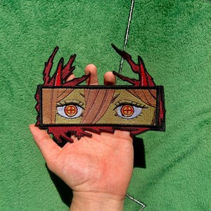 Peut inclure: Écusson brodé représentant des yeux de style anime avec des iris rouges et un motif en croix. L'écusson a une forme rectangulaire avec une bordure marron et des accents rouges en forme de flamme. Fond vert.