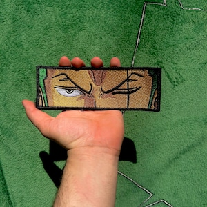 Peut inclure: Écusson brodé représentant un gros plan des yeux d'un personnage, avec une expression sévère. L'écusson est de couleur or, vert et noir. Il est rectangulaire avec une bordure noire. Le fond est un tissu vert texturé.
