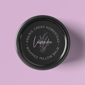 May include: A black jar lid with the words "Prairie Creek Homestead" and "Whipped Tallow Balm" encircling the center. The word "Lavender" is written in cursive with a lavender sprig illustration in the center. The background is a light purple.